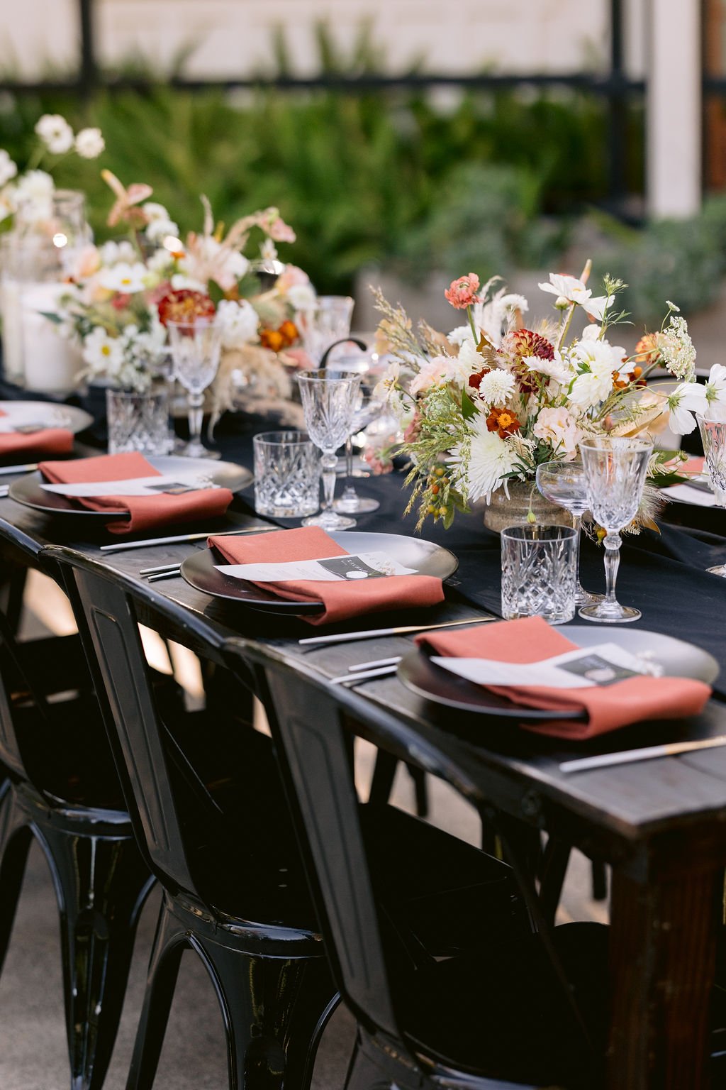 A long dining table set for an event with black chairs, elegantly folded orange napkins, crystal glassware, and floral centerpieces featuring a mix of white, yellow, and red flowers.