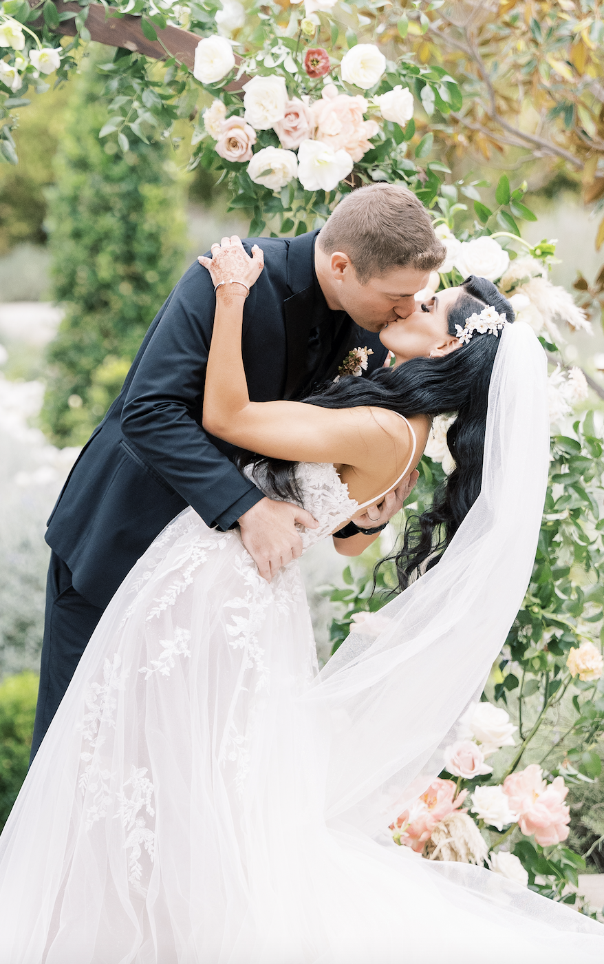 A bride and groom share a kiss while standing under a floral arch in an outdoor garden setting. The bride is wearing a white dress with a veil, and the groom is in a black suit.