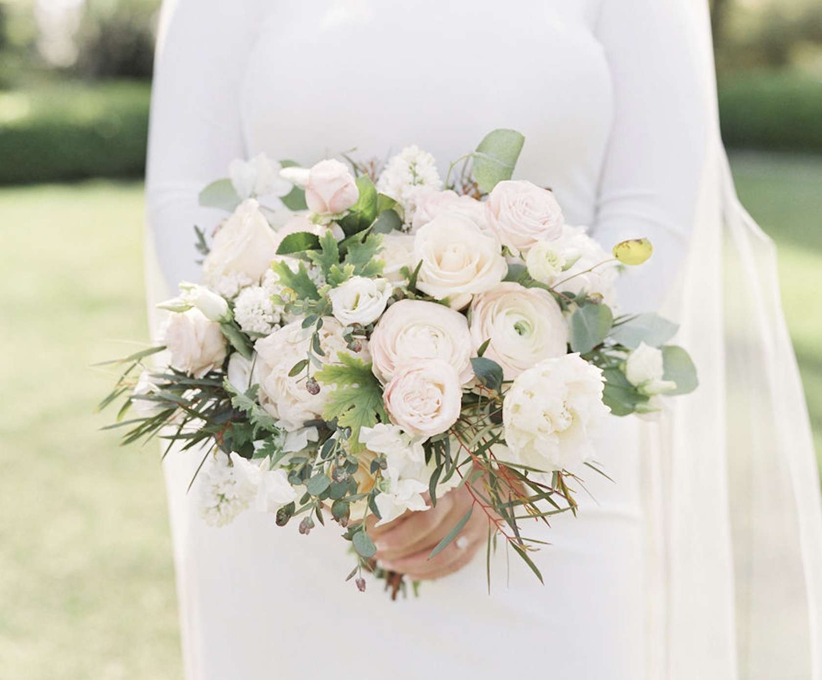 A person in a white outfit holds a bouquet of white and pale pink flowers with greenery. The background is a garden or outdoor setting.