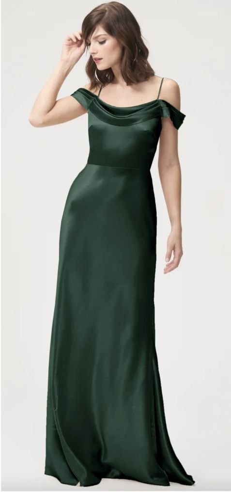 A woman stands wearing an elegant, floor-length, dark green satin off-shoulder gown with spaghetti straps, looking down and touching her hair.