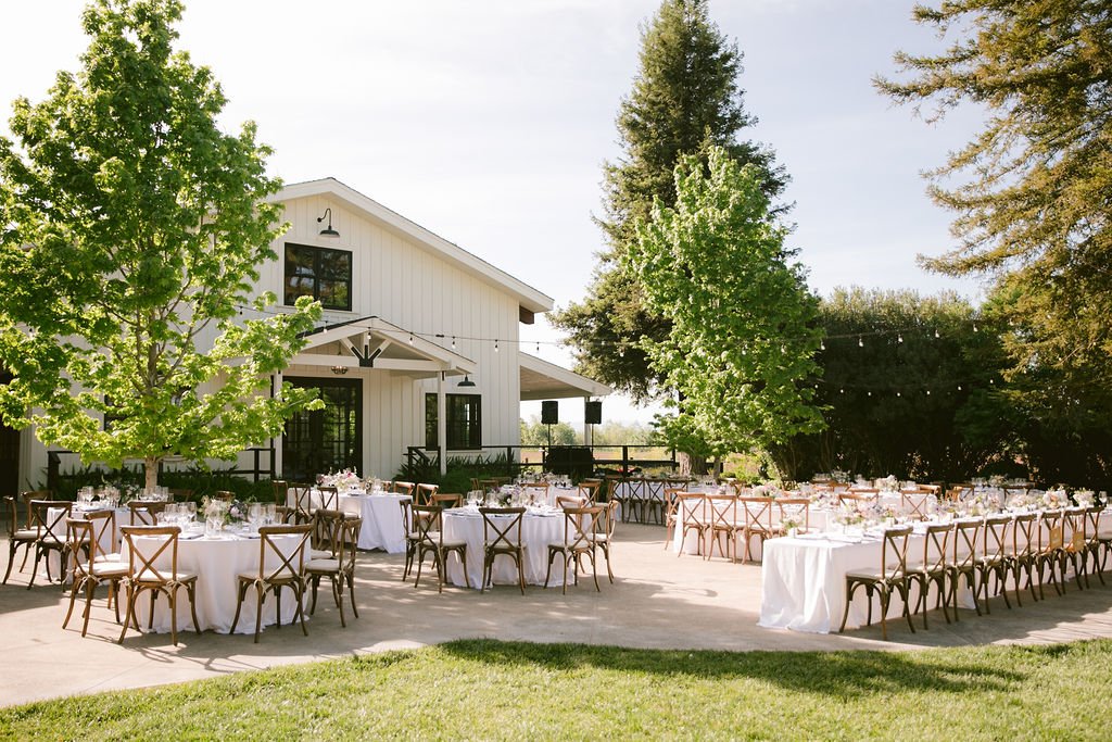 Outdoor wedding reception setup with round and rectangular tables covered in white linens, arranged in front of a light-colored barn. String lights are hung between trees, and green lawn surrounds the area.