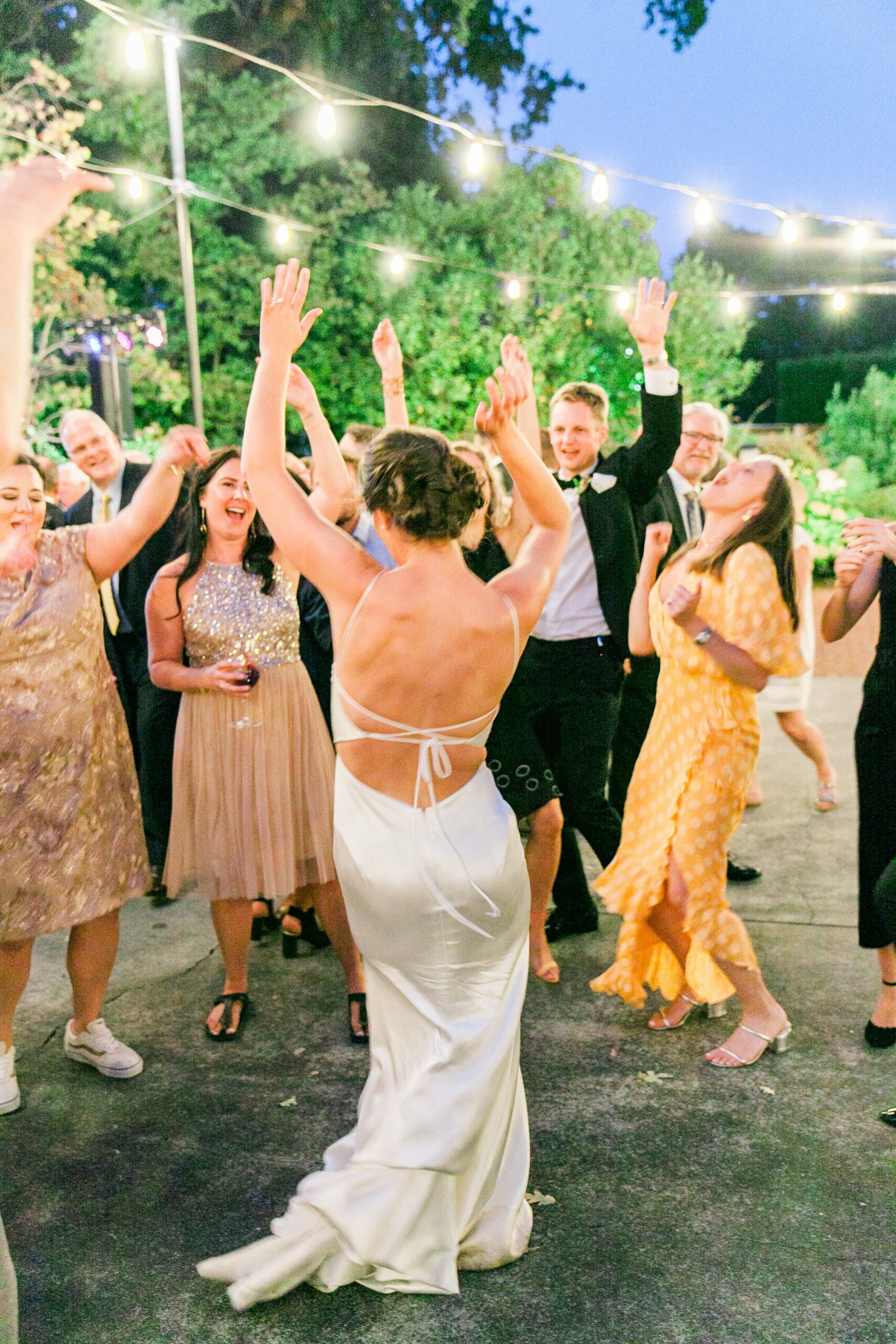 A group of people dressed in semi-formal attire are dancing and celebrating outdoors under string lights at night.