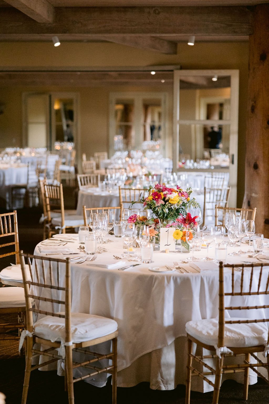 A round table set for a formal event with white tablecloth, glassware, silverware, and a floral centerpiece with a table number "1." Gold chairs surround the table in a dimly lit room.