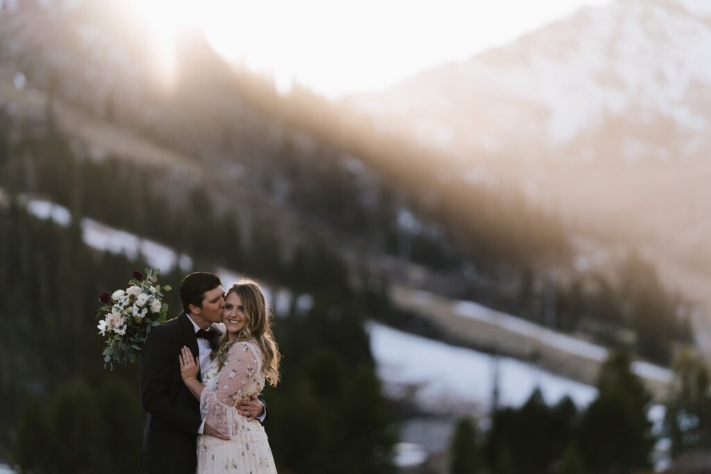 A couple embraces in front of a mountainous landscape with snow patches. The man is in a black suit, and the woman is in a light dress, holding a bouquet of flowers. Sunlight filters through the scene.