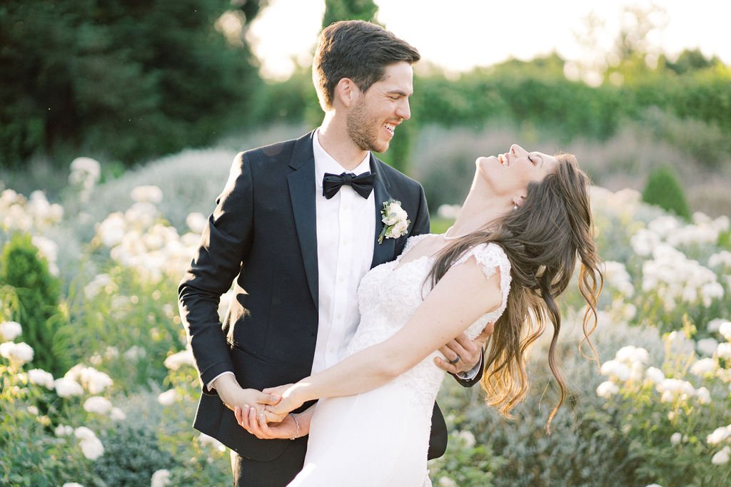 A man in a black suit and bow tie holds a woman in a white gown as they smile at each other outdoors, surrounded by greenery and white flowers.