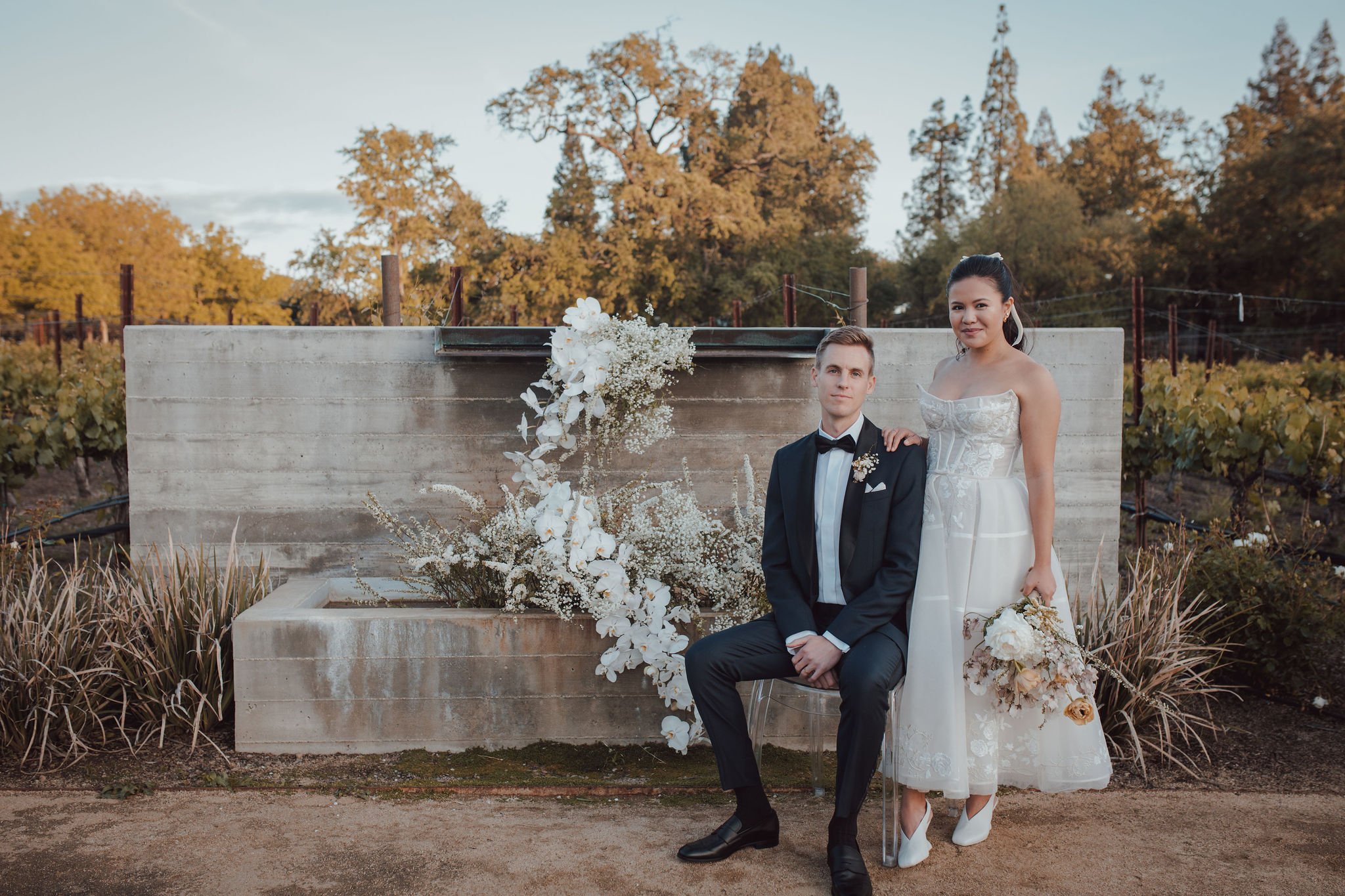 A couple in wedding attire poses outdoors by a concrete structure adorned with white flowers. The groom is seated, and the bride stands next to him holding a bouquet. Trees and vineyard are in the background.