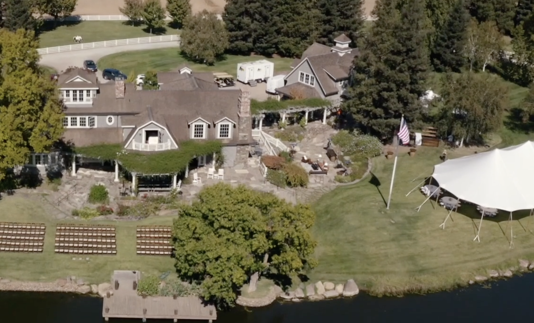 Aerial view of a large house with surrounding greenery, an American flag, a white tent, a landscaped yard, and a nearby body of water.