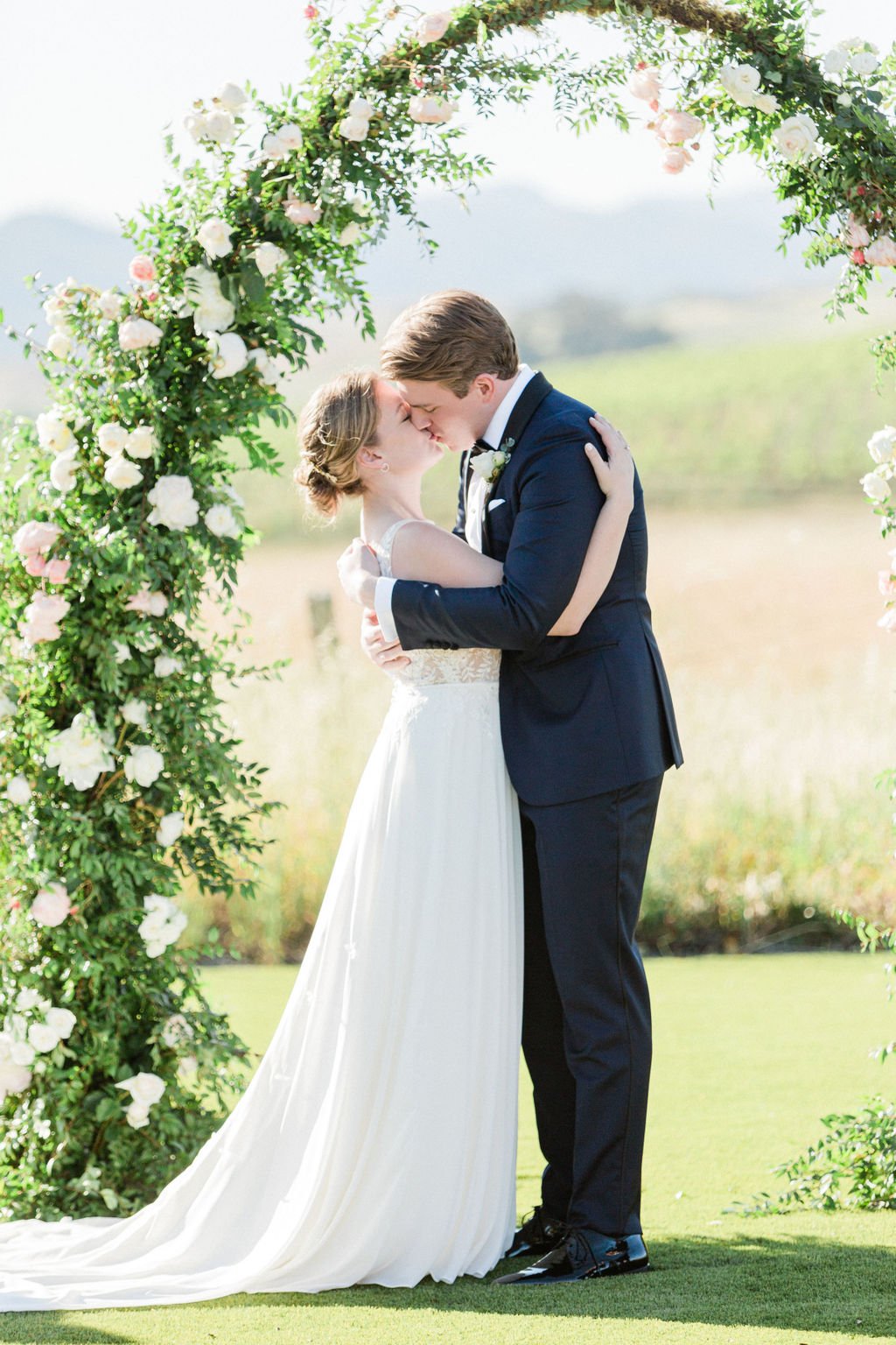 A couple is kissing under a floral arch outdoors. The woman is in a white wedding dress, and the man is in a dark suit. The background shows a grassy field and distant hills.