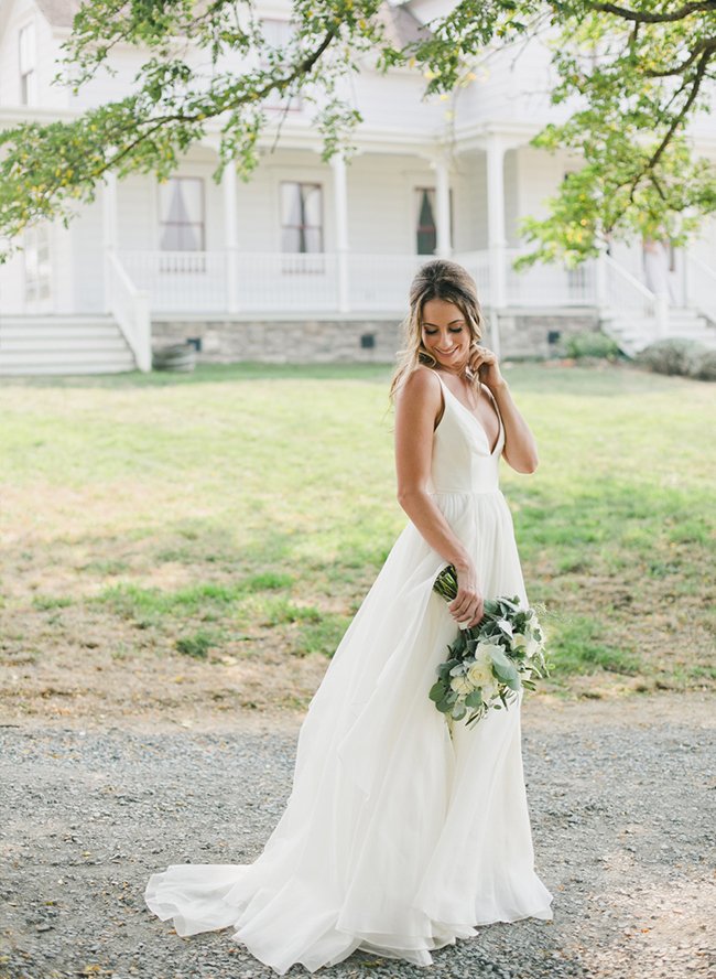 A bride in a white gown holds a floral bouquet while standing outdoors in front of a large white house with a porch.