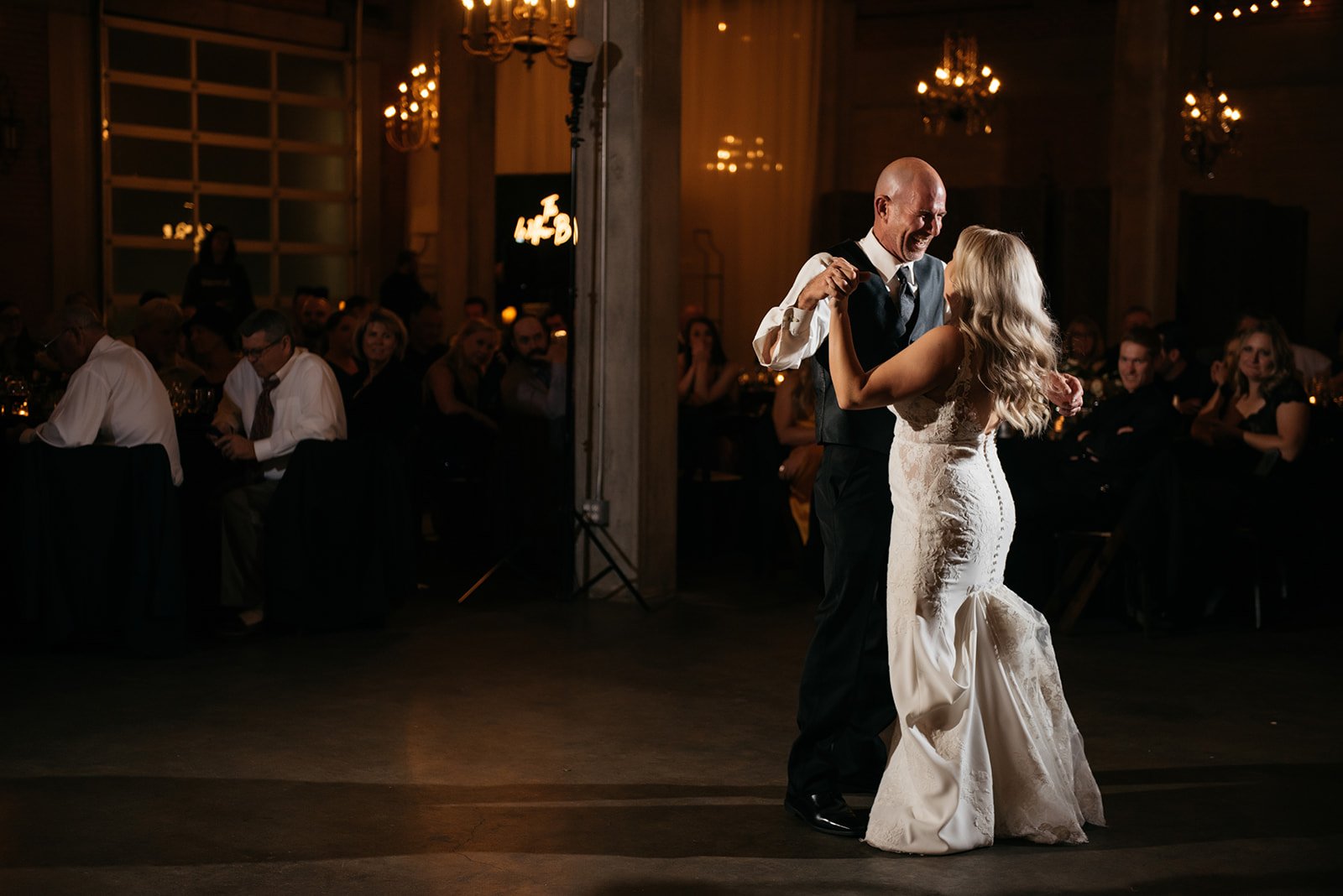 A couple dances together in a dimly lit room with chandeliers and people seated at tables in the background.