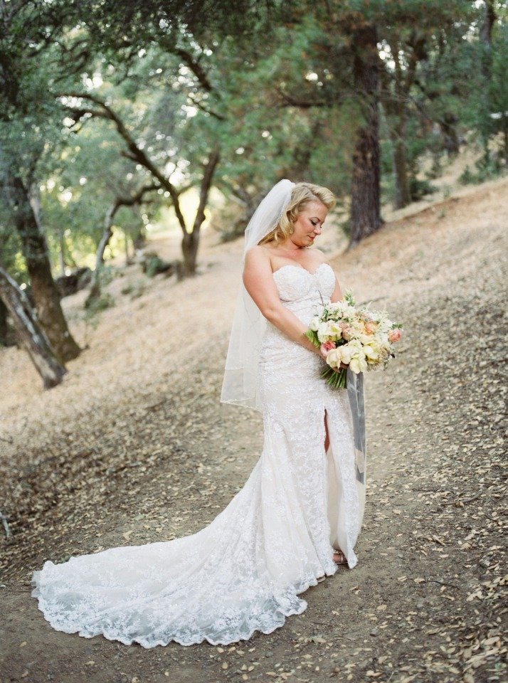 A bride in a white gown and veil holds a bouquet of flowers while standing on a wooded path covered in leaves.