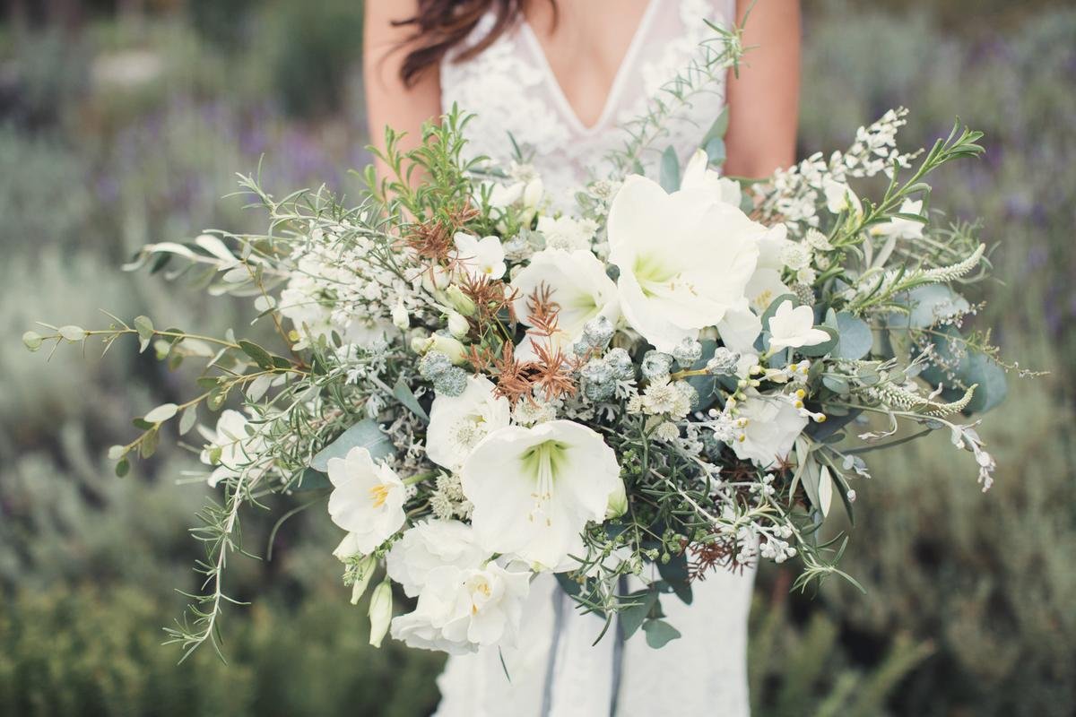 A woman in a white dress holds a large bouquet of white flowers and greenery against a blurred outdoor background.