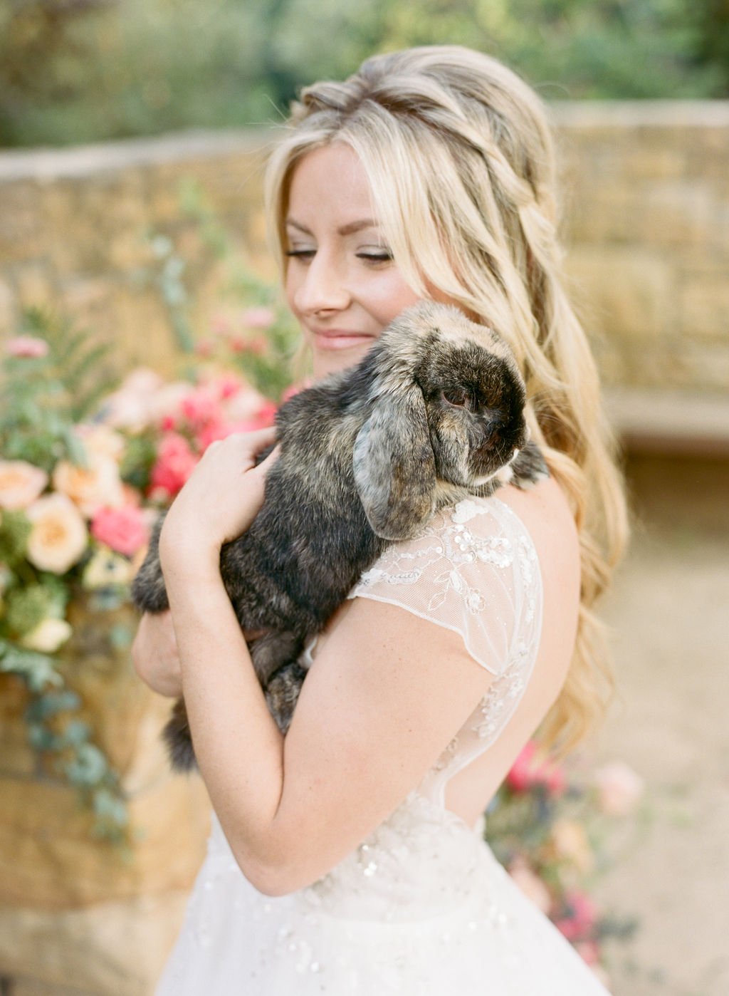 A person in a white dress holding a rabbit, standing in front of a stone wall and floral arrangement.