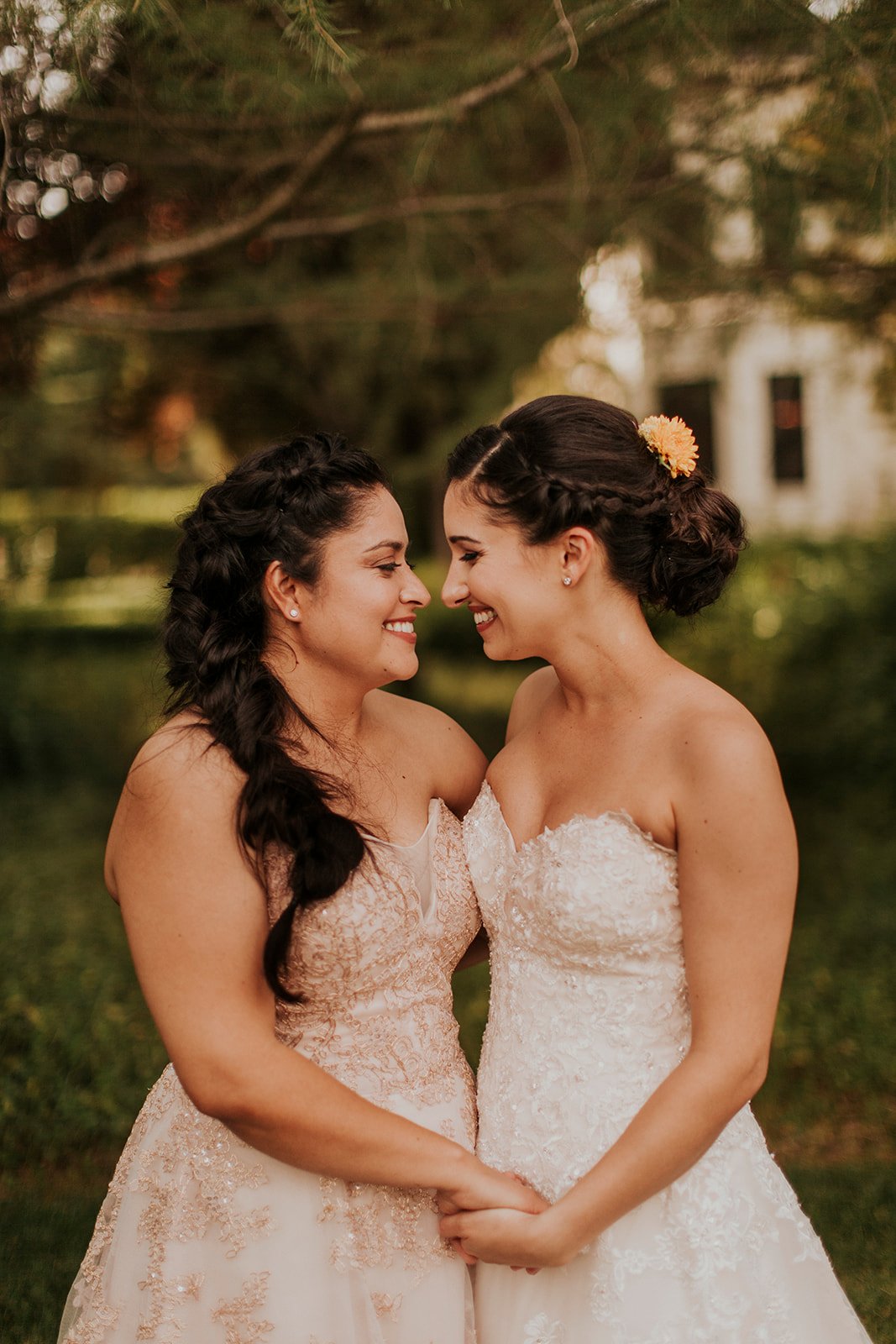 Two women in wedding dresses stand outdoors, holding hands, and smiling at each other with green foliage in the background.