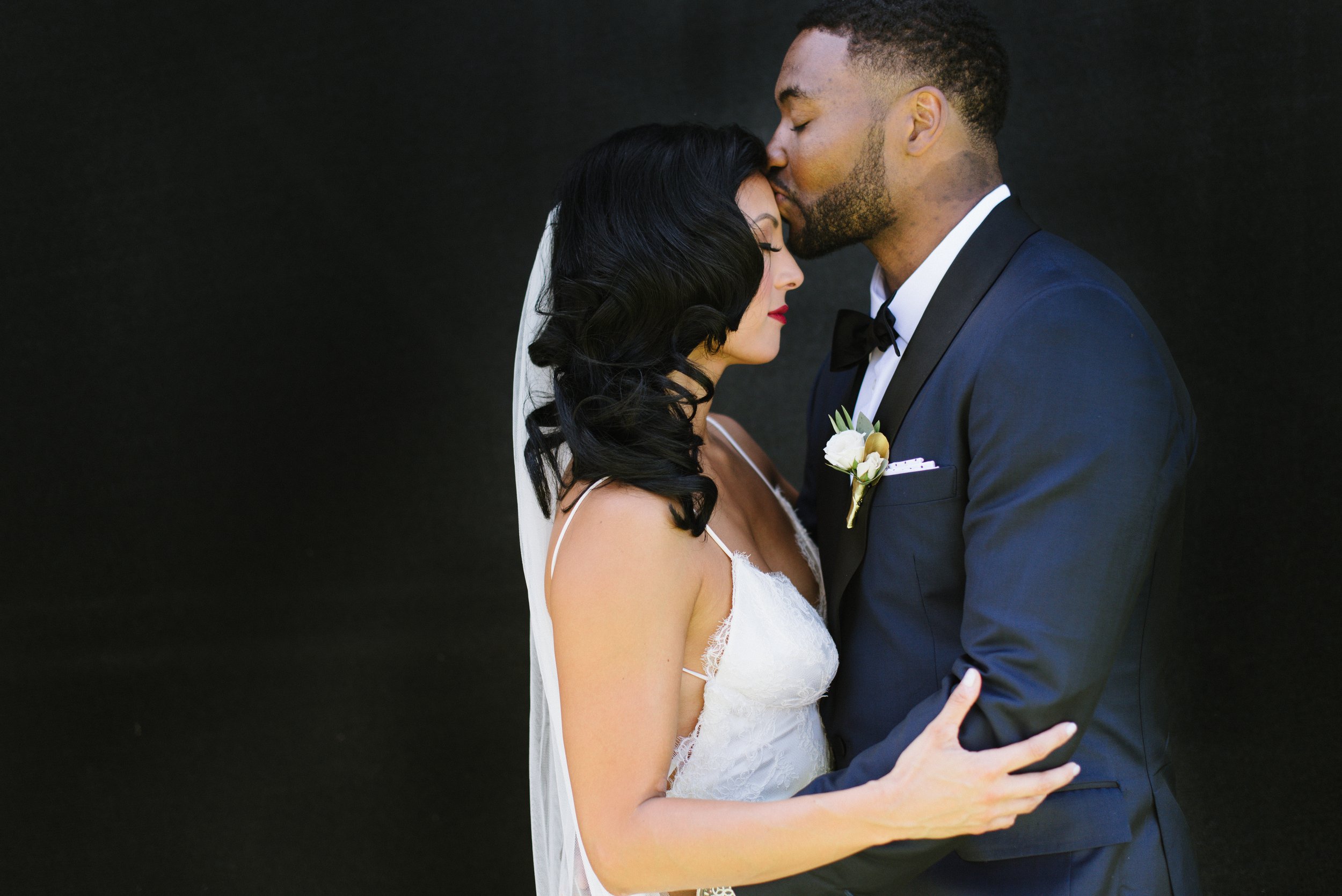 A groom kisses the bride's forehead while embracing her. The bride wears a white dress and veil, and the groom wears a dark suit and bow tie.