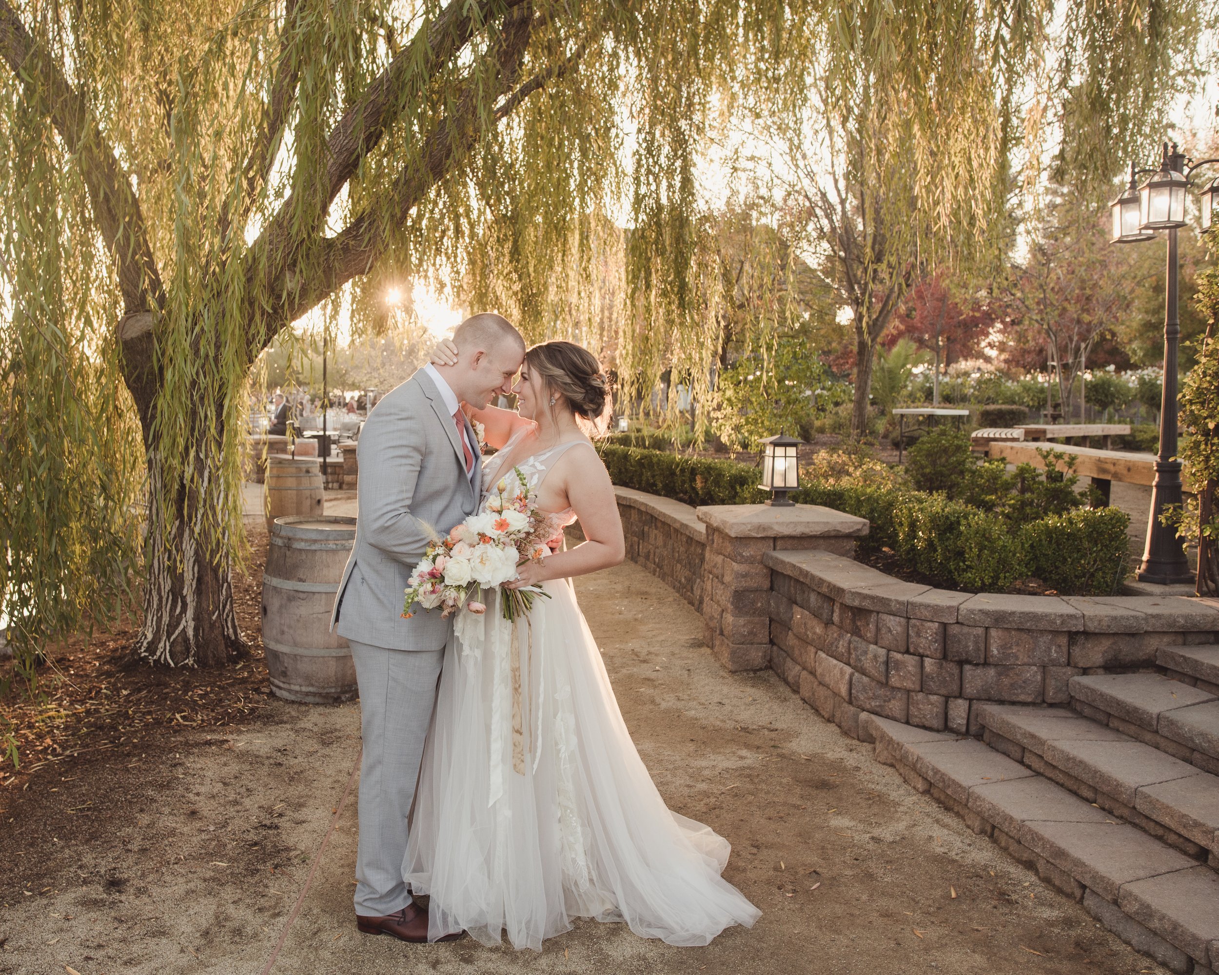 A bride and groom share an intimate moment under a tree in a park-like setting, with the bride holding a bouquet and wearing a white gown, and the groom in a light gray suit.