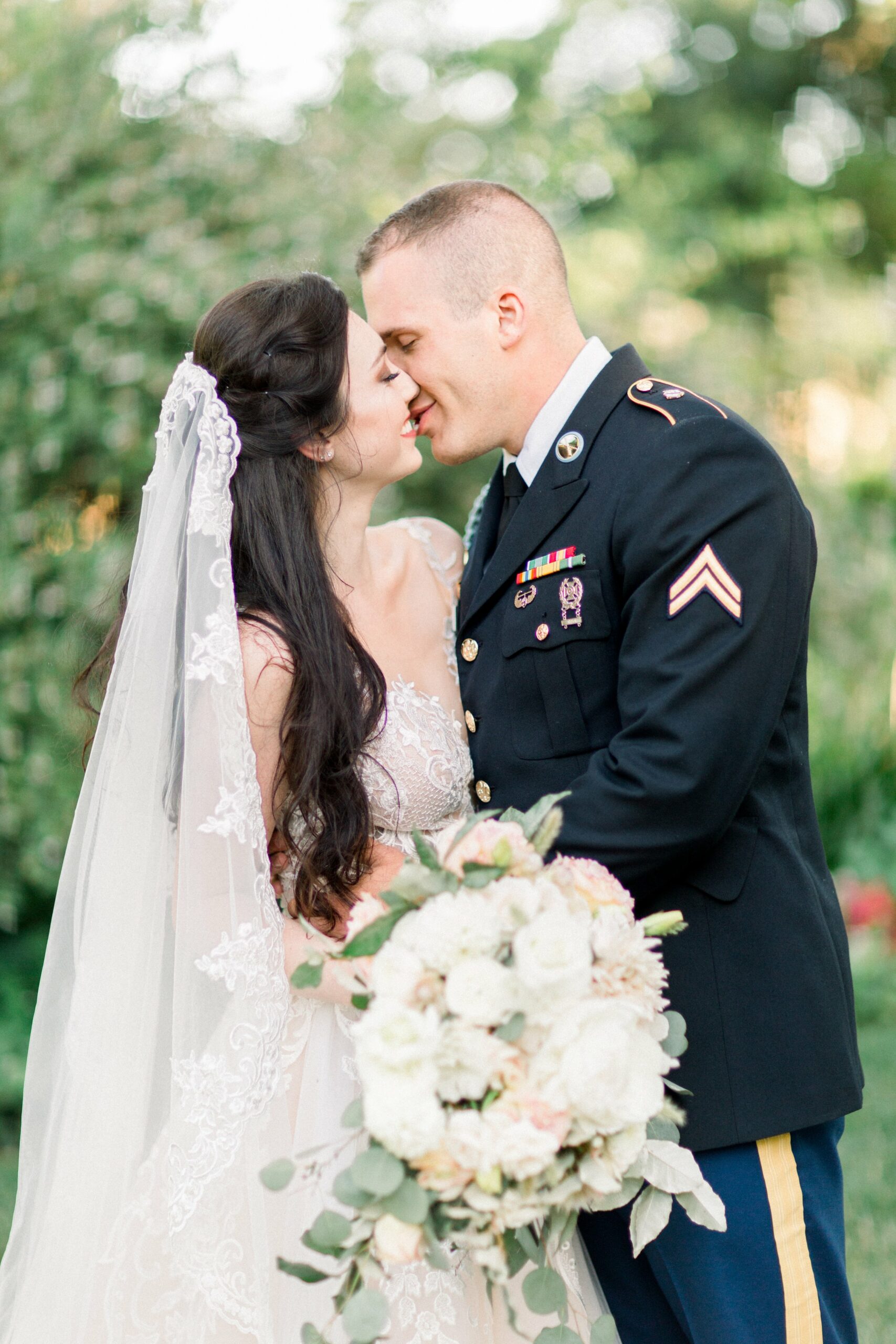 A bride in a white dress and veil holds a bouquet of flowers while smiling closely with a groom in a military uniform outdoors.