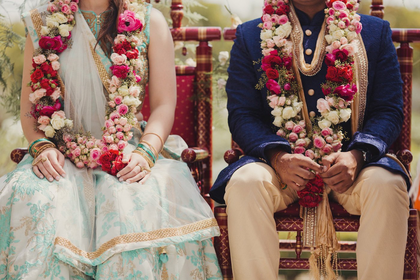 A couple, adorned with large floral garlands, sits side by side on patterned chairs, dressed in traditional attire.