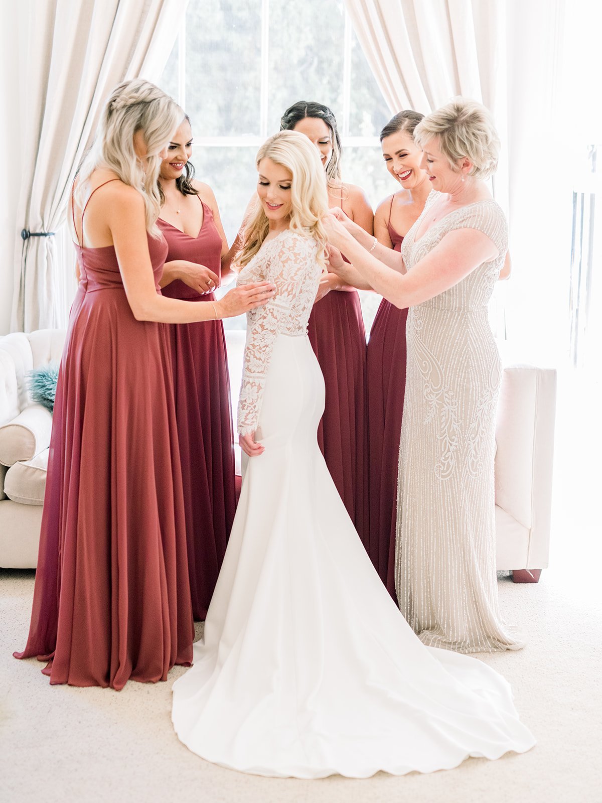 A bride in a white dress is being assisted by four women in maroon and beige dresses in a well-lit room.