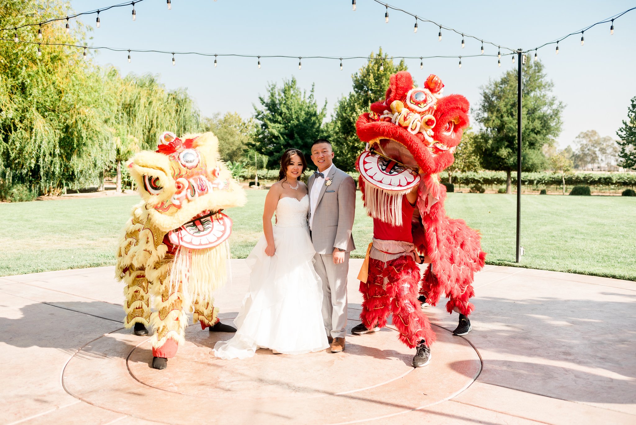 A bride and groom stand outdoors under string lights, flanked by two performers in colorful Chinese lion dance costumes.