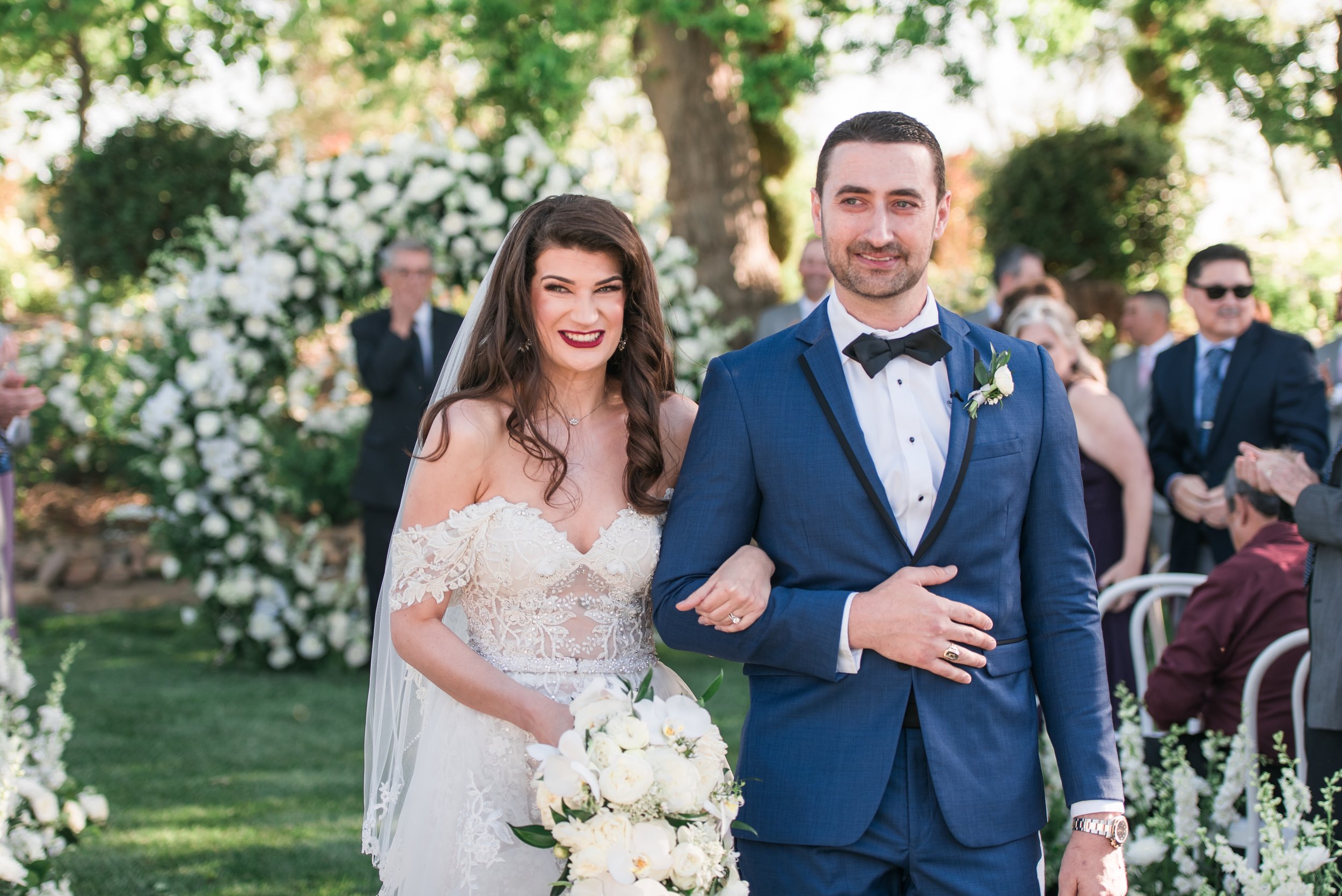 A bride in a white gown and veil and a groom in a blue suit with a boutonniere stand together smiling outdoors, with guests and floral decorations in the background.