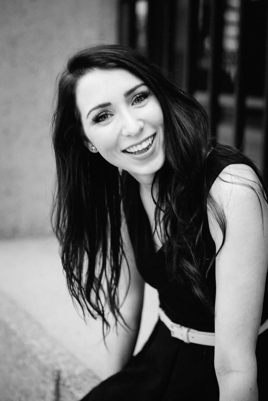 Black and white photo of a woman with long dark hair, smiling broadly, wearing a sleeveless dark top and a light-colored belt, sitting outdoors.