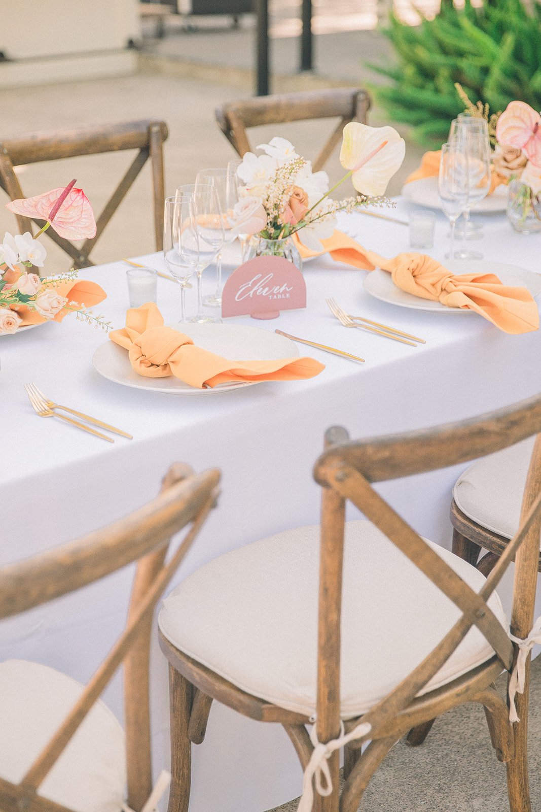 A dining table set outdoors with wooden chairs, white tablecloth, gold cutlery, orange napkins, and floral centerpieces.