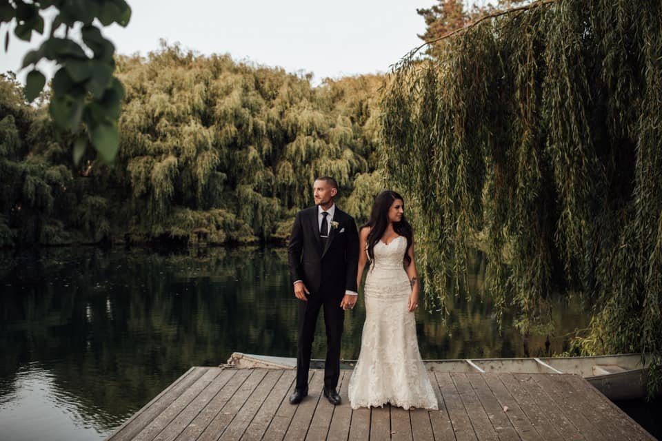 A bride and groom stand on a wooden dock by a serene lake with lush greenery in the background.