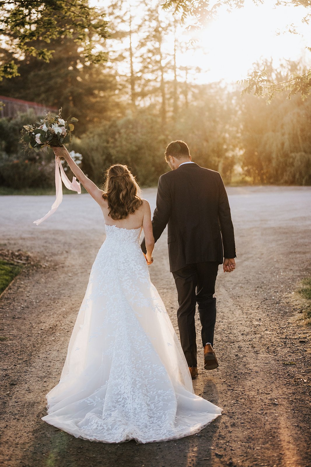 A bride in a white gown and a groom in a dark suit walk hand in hand on a sunlit path, with the bride raising a bouquet in her left hand.