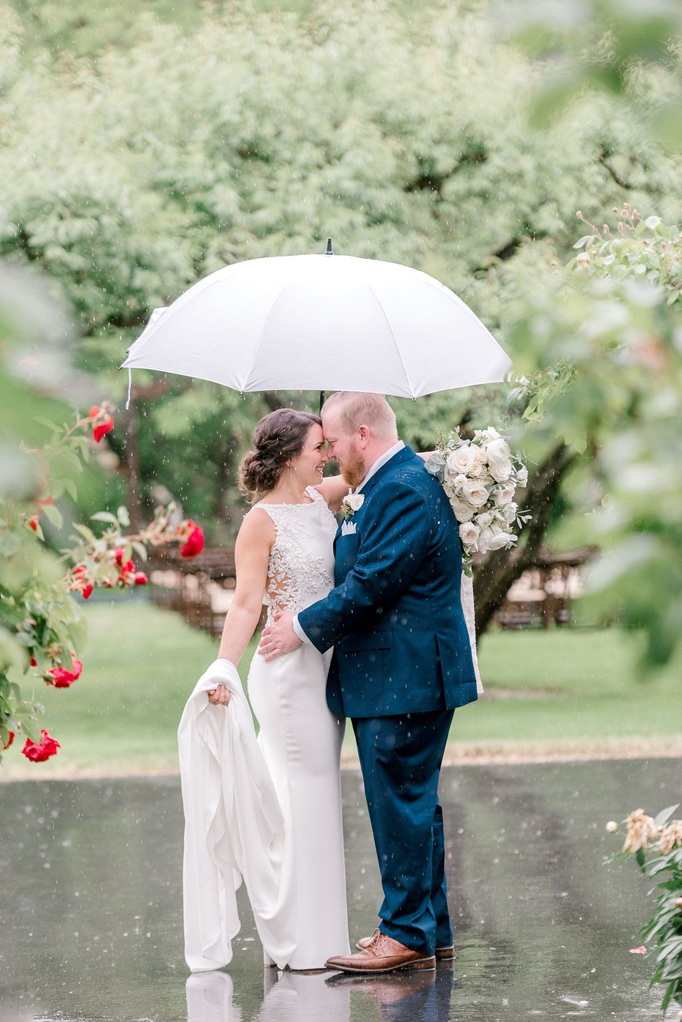 A bride and groom stand close under a large white umbrella in a garden, surrounded by greenery and red flowers. The bride holds her dress while the groom holds the bouquet. It is raining lightly.