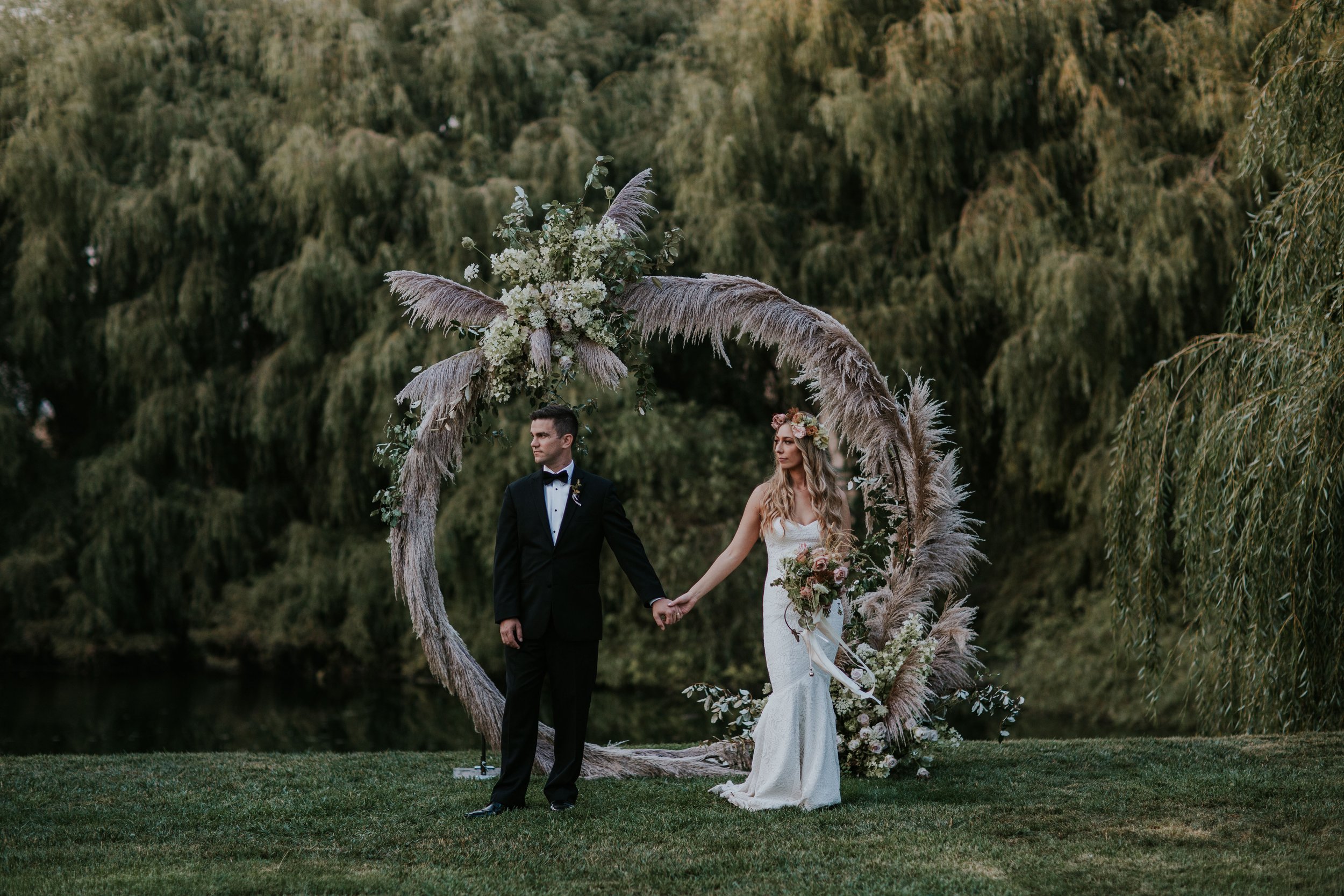 A couple stands holding hands in front of a circular floral arch outdoors, with the groom in a black suit and the bride in a white dress holding a bouquet.