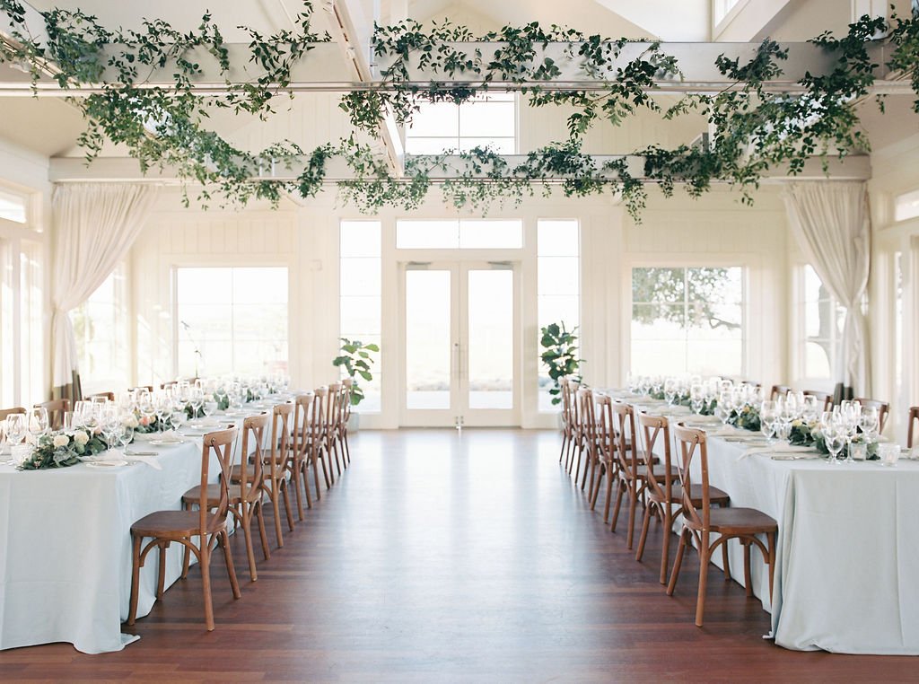 A bright banquet hall with wooden flooring features two long, white-clothed tables and wooden chairs. Ceiling greenery decorates the space.