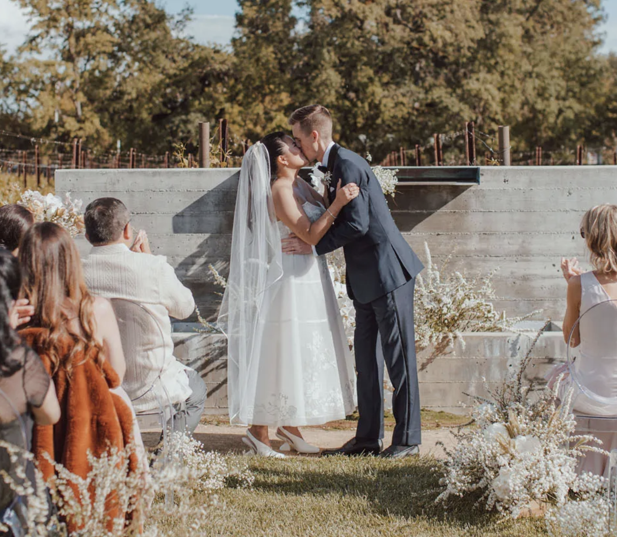 A bride and groom kiss at an outdoor wedding ceremony, with seated guests watching and a natural backdrop of trees and flowers.