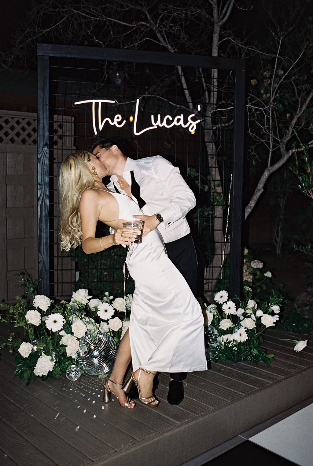 A couple dressed in formal attire kisses in front of a neon sign that reads "The Lucas'." They are surrounded by white flowers and greenery. The woman holds a drink, and they both appear happy.