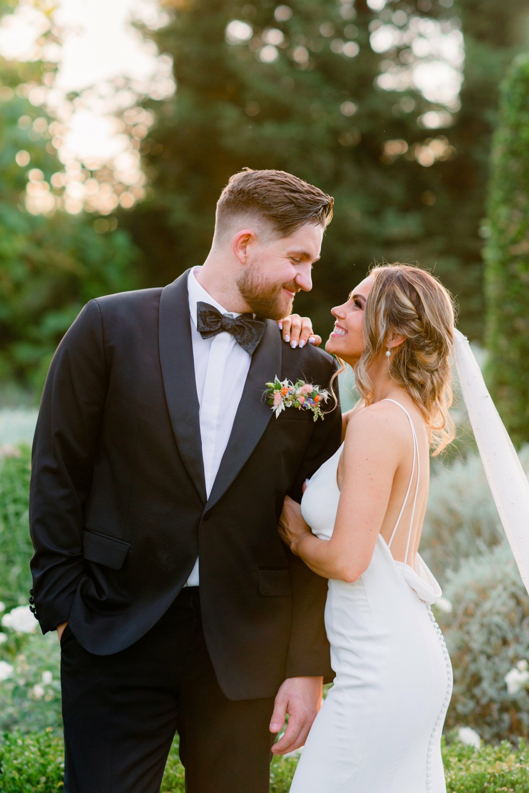 A bride in a white dress and veil leans affectionately on a smiling groom in a black suit with a boutonnière, standing in an outdoor garden.