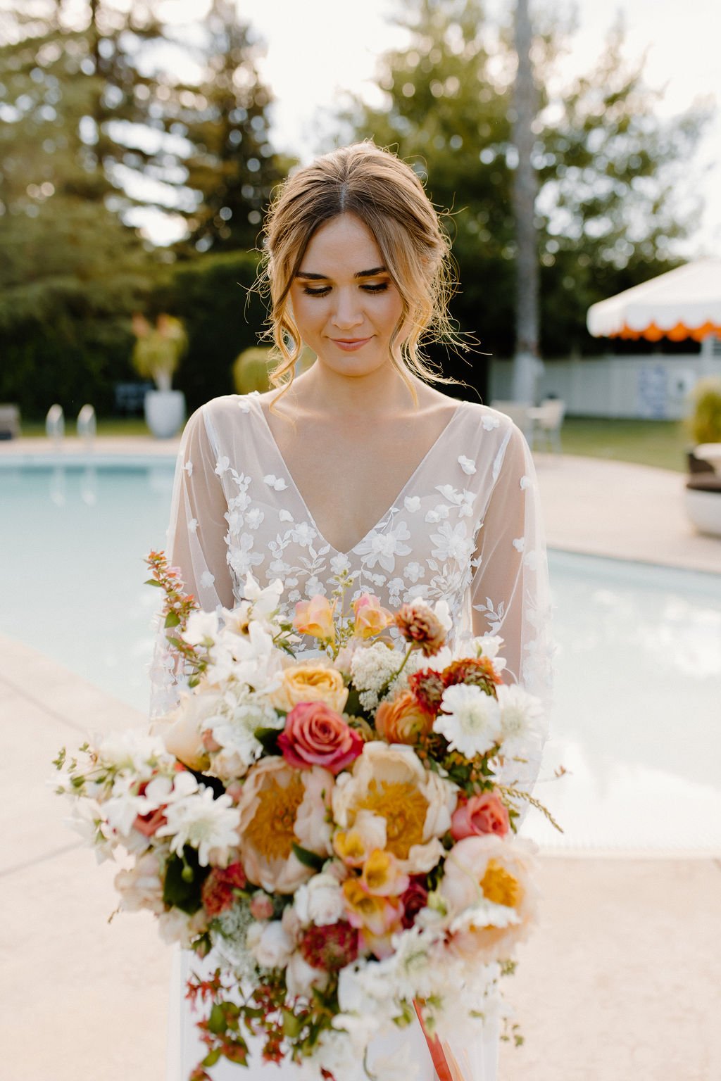 A woman in a white dress holding a large bouquet of multicolored flowers stands by a pool in an outdoor setting with trees and a tent in the background.