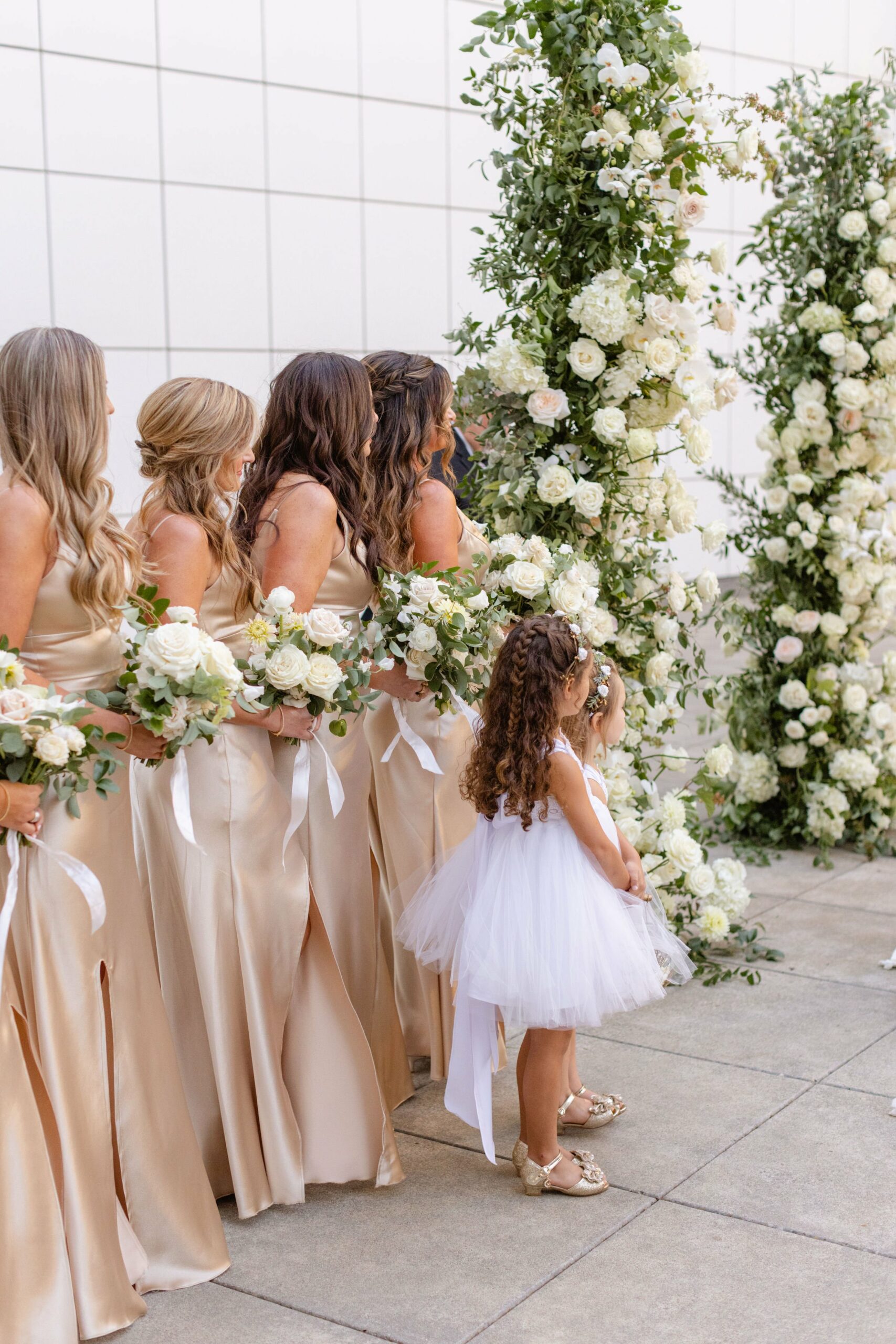 Bridesmaids in gold dresses and a flower girl in a white dress stand in front of a floral arch made of white roses and greenery during a wedding ceremony.