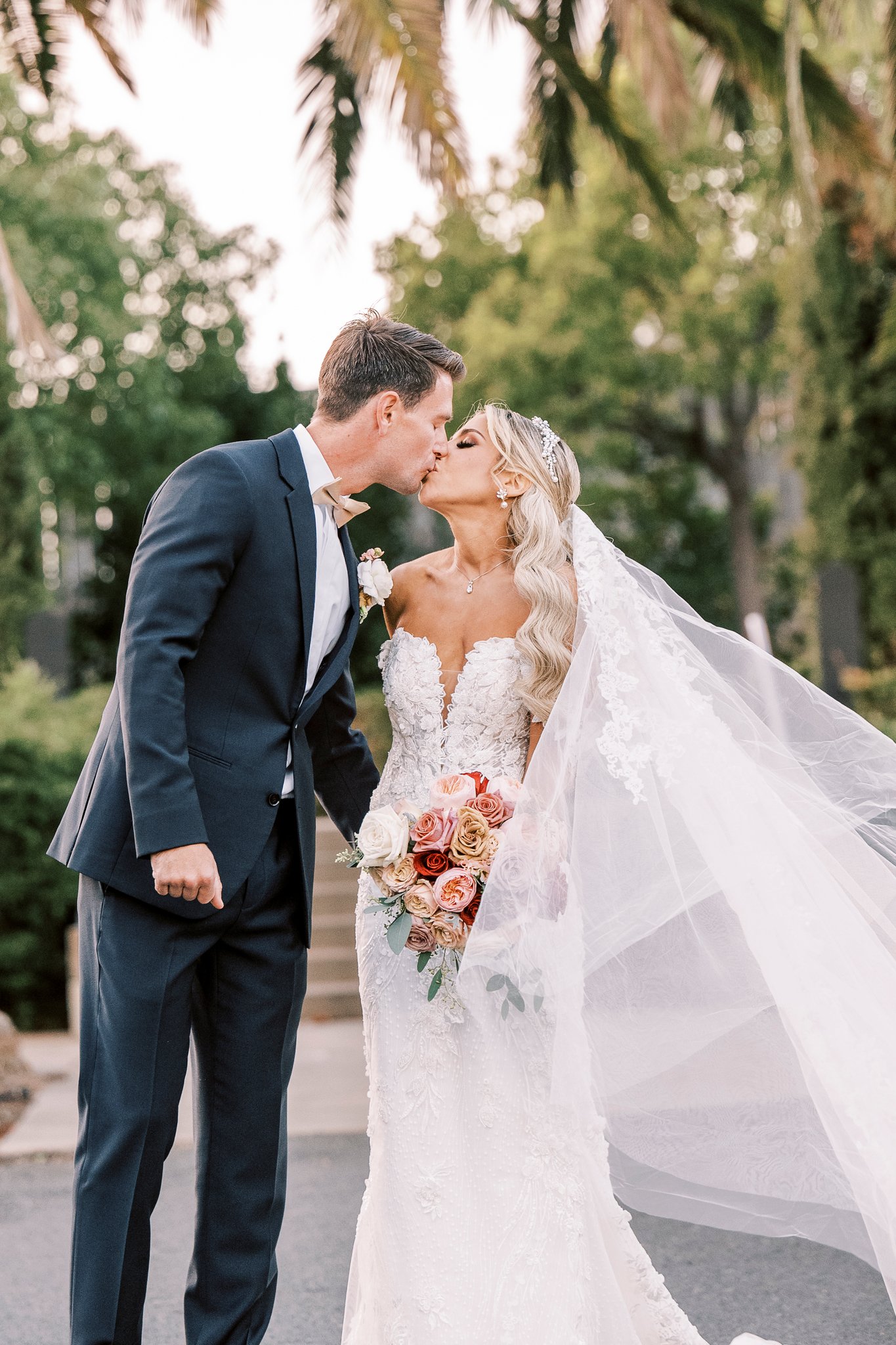 A bride in a white gown and veil holds a bouquet of flowers and kisses a groom in a dark suit outdoors with trees in the background.