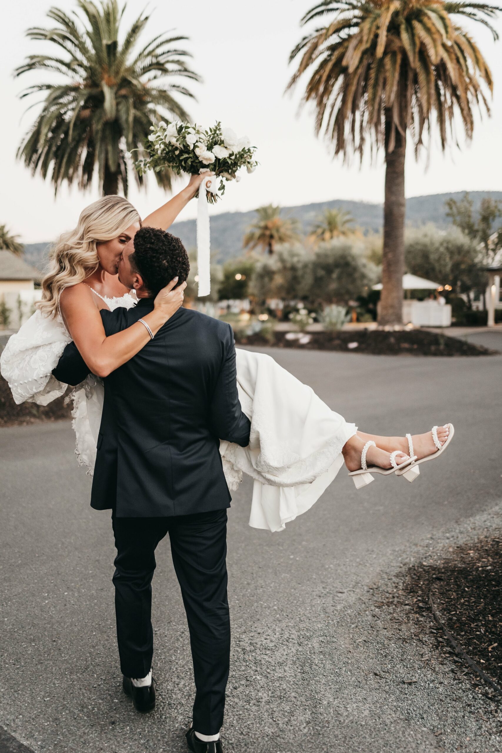 A man in a suit carries a woman in a wedding gown on a tree-lined street, while she holds a bouquet aloft.