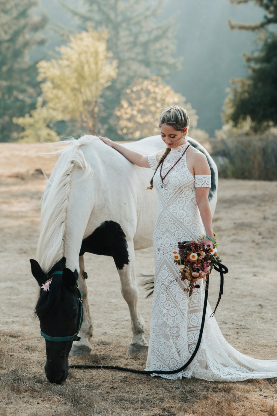 A woman in a white lace dress stands beside a white and black horse, holding a bouquet of flowers, in an outdoor setting with trees in the background.