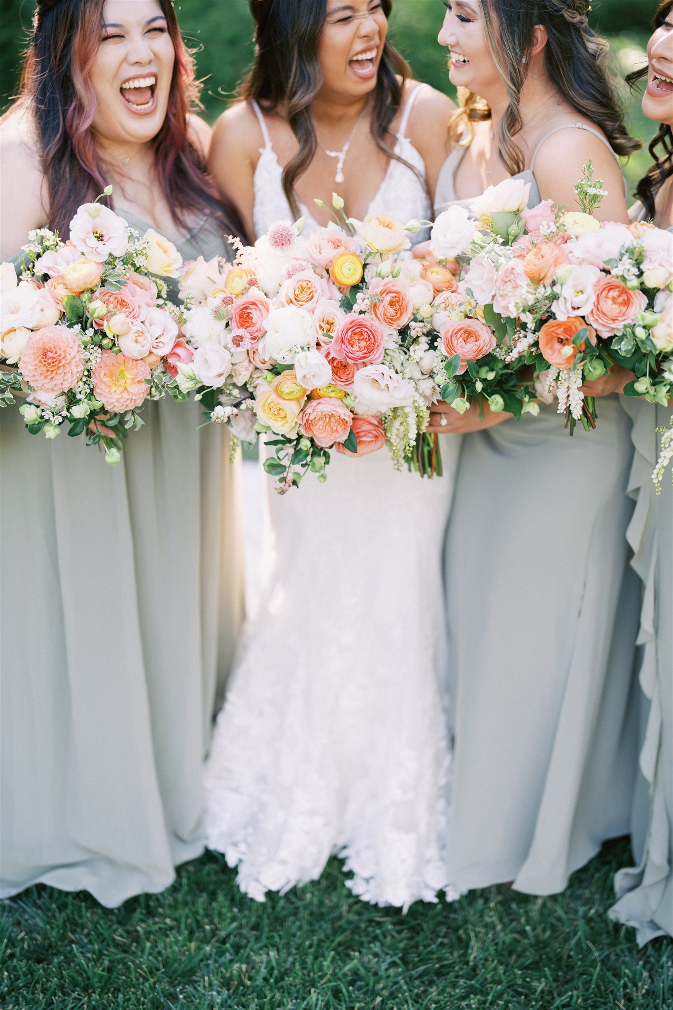 Four women in dresses, three in sage green and one in white, hold large bouquets of flowers and laugh together outdoors.