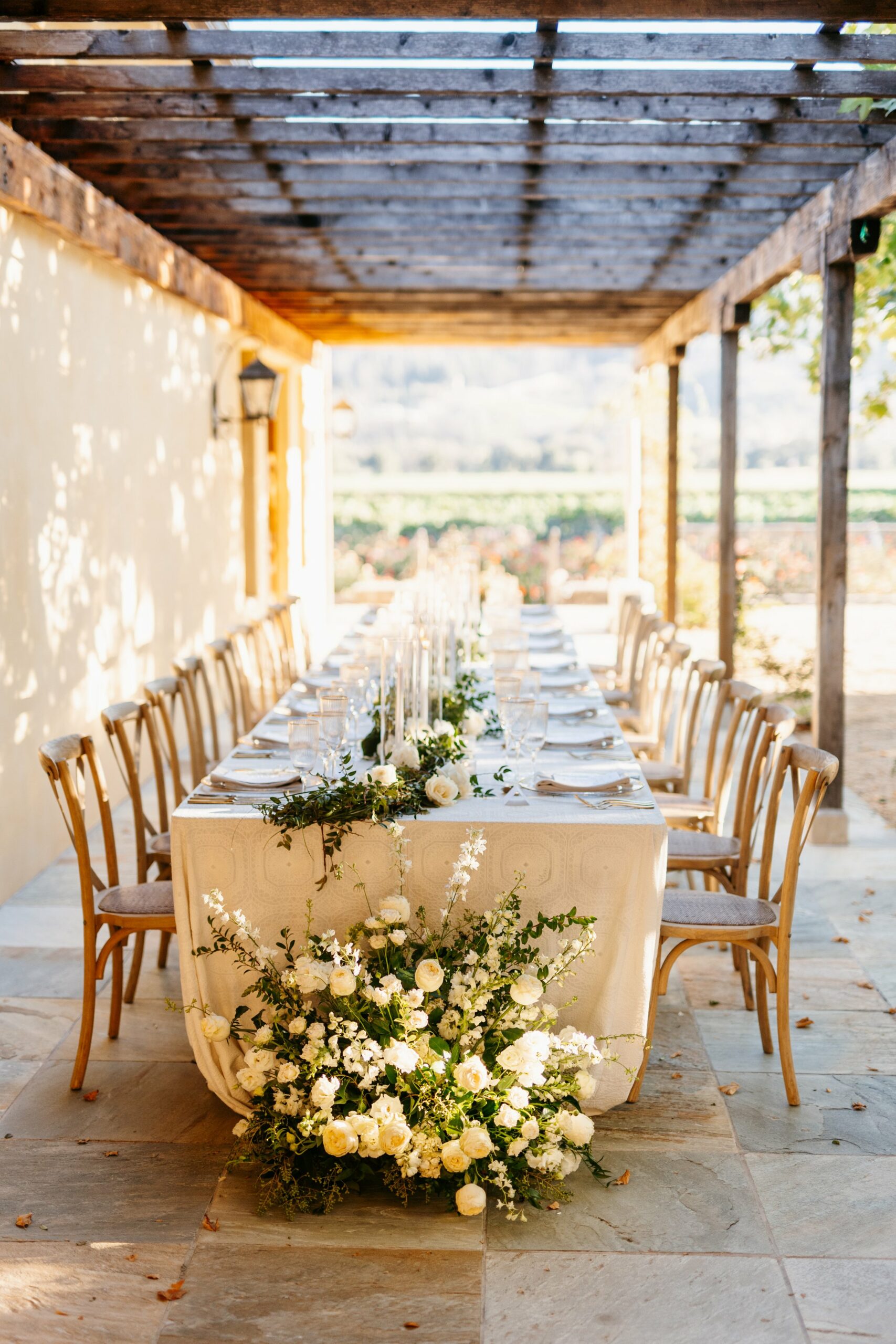 A long dining table set for an outdoor event, adorned with white flowers and greenery, positioned under a wooden pergola.