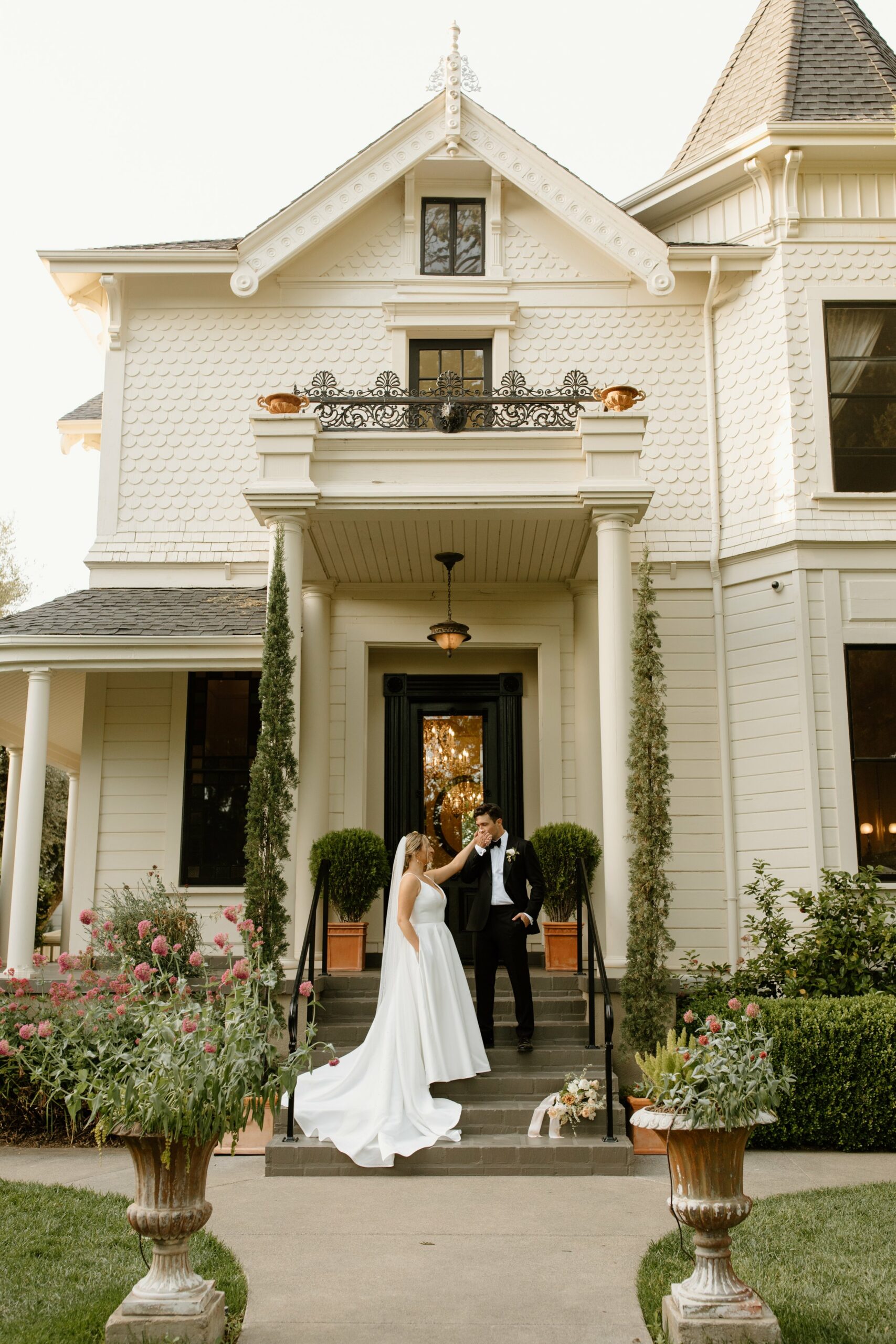 A bride and groom stand on the stairs of a large, white, Victorian-style house. The bride is in a white dress, and the groom is in a black suit, surrounded by green plants and flowers.