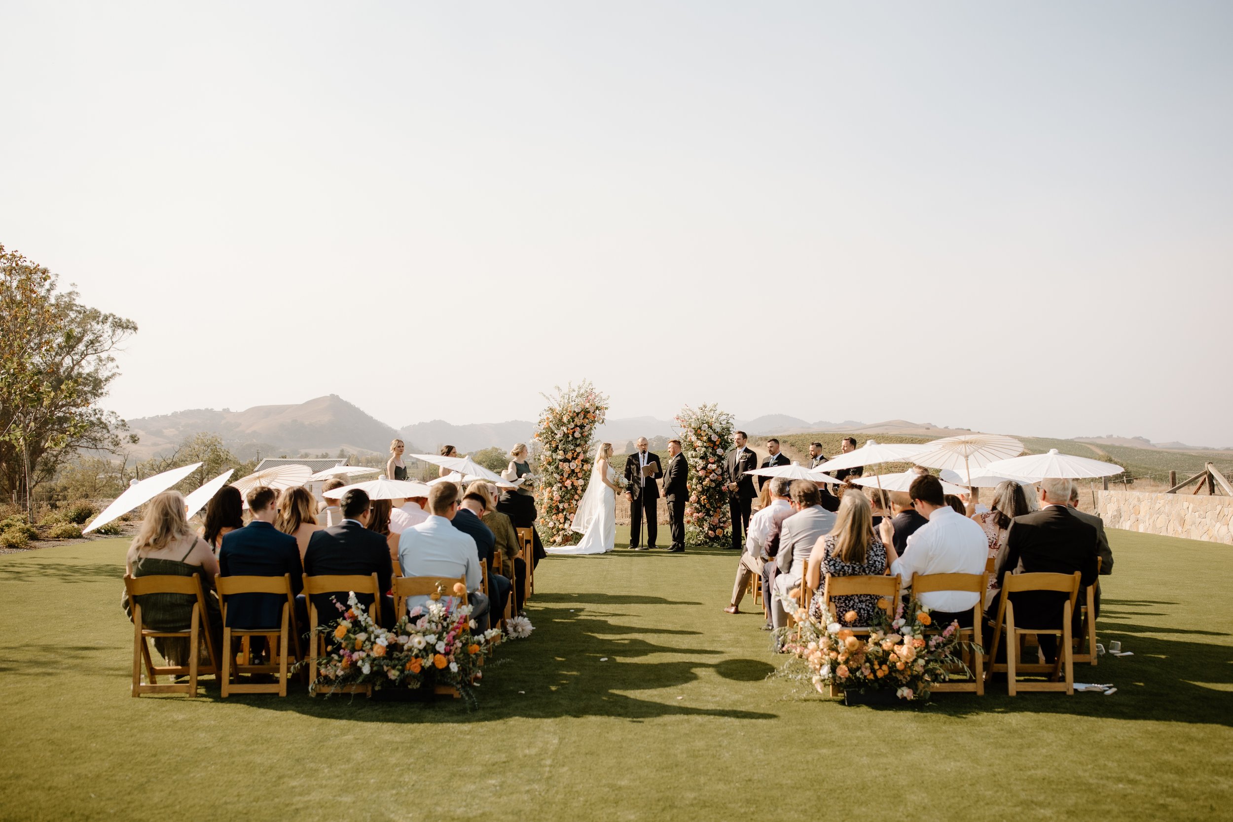 Outdoor wedding ceremony with a small crowd seated on either side of an aisle adorned with flowers. The couple stands in front of a floral arch, and a celebrant is officiating. Hills are visible in the background.