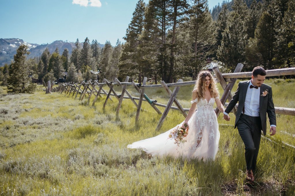 A couple in wedding attire walks hand-in-hand through a grassy field with a wooden fence and trees in the background. The bride holds a bouquet of flowers.