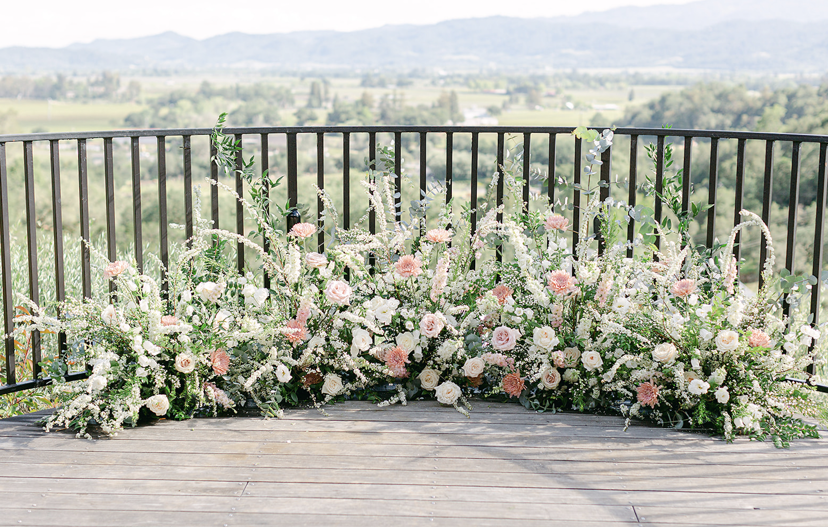 An outdoor area overlooks a scenic landscape. The foreground features a decorative floral arrangement with a variety of white and pink flowers situated against a black metal railing.