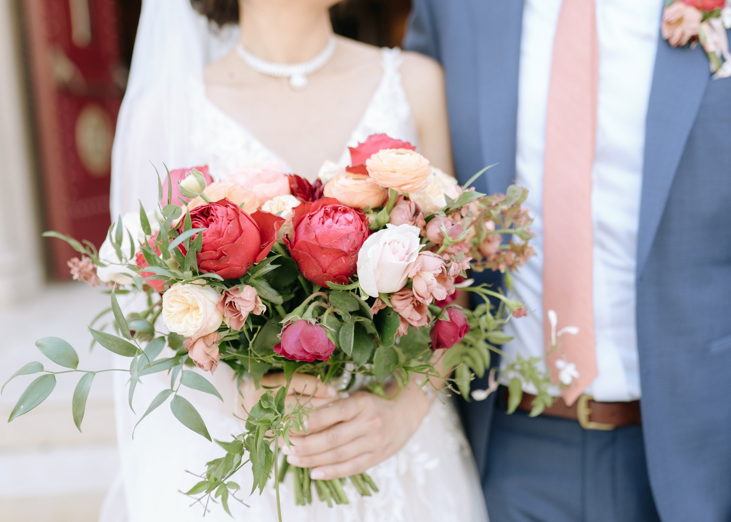 A bride in a white dress holds a bouquet of red, pink, and peach flowers, standing beside a groom in a blue suit and pink tie.