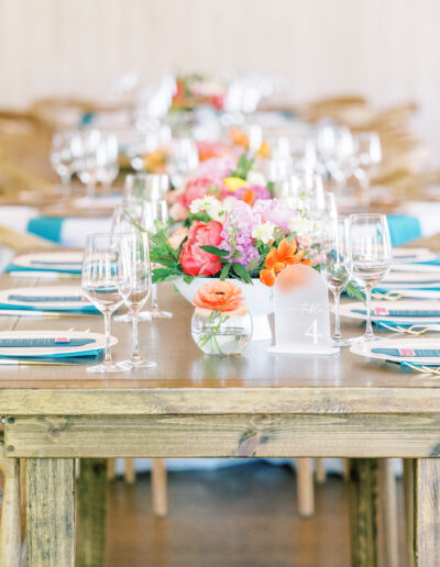 A wooden dining table is set with glassware, plates, menus, teal napkins, and colorful floral centerpieces, surrounded by wooden chairs with teal sashes.