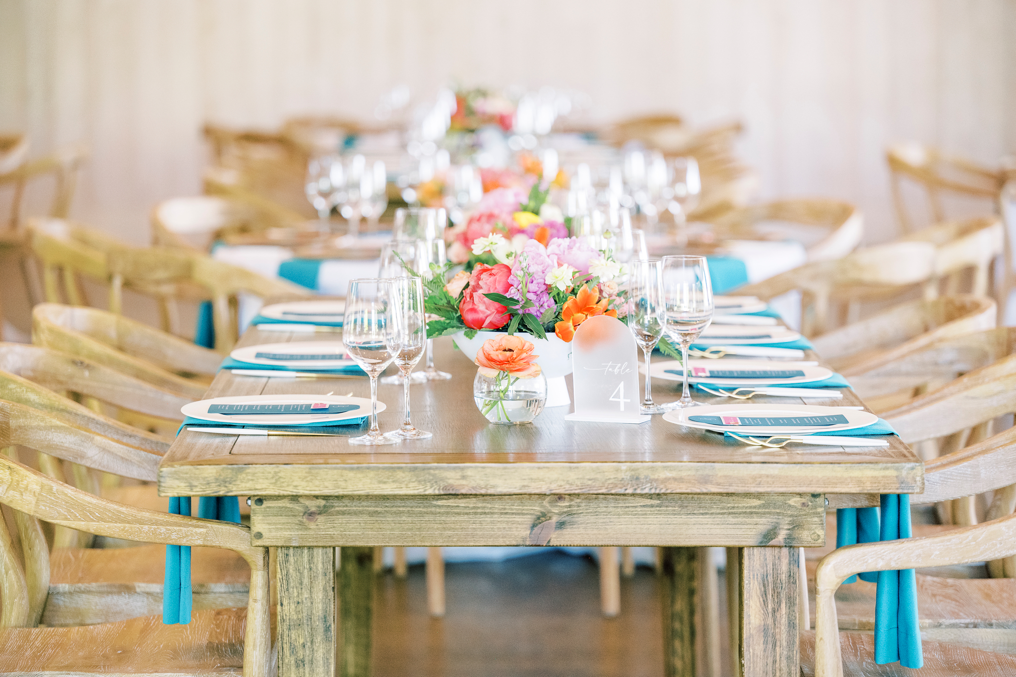A wooden dining table is set with glassware, plates, menus, teal napkins, and colorful floral centerpieces, surrounded by wooden chairs with teal sashes.