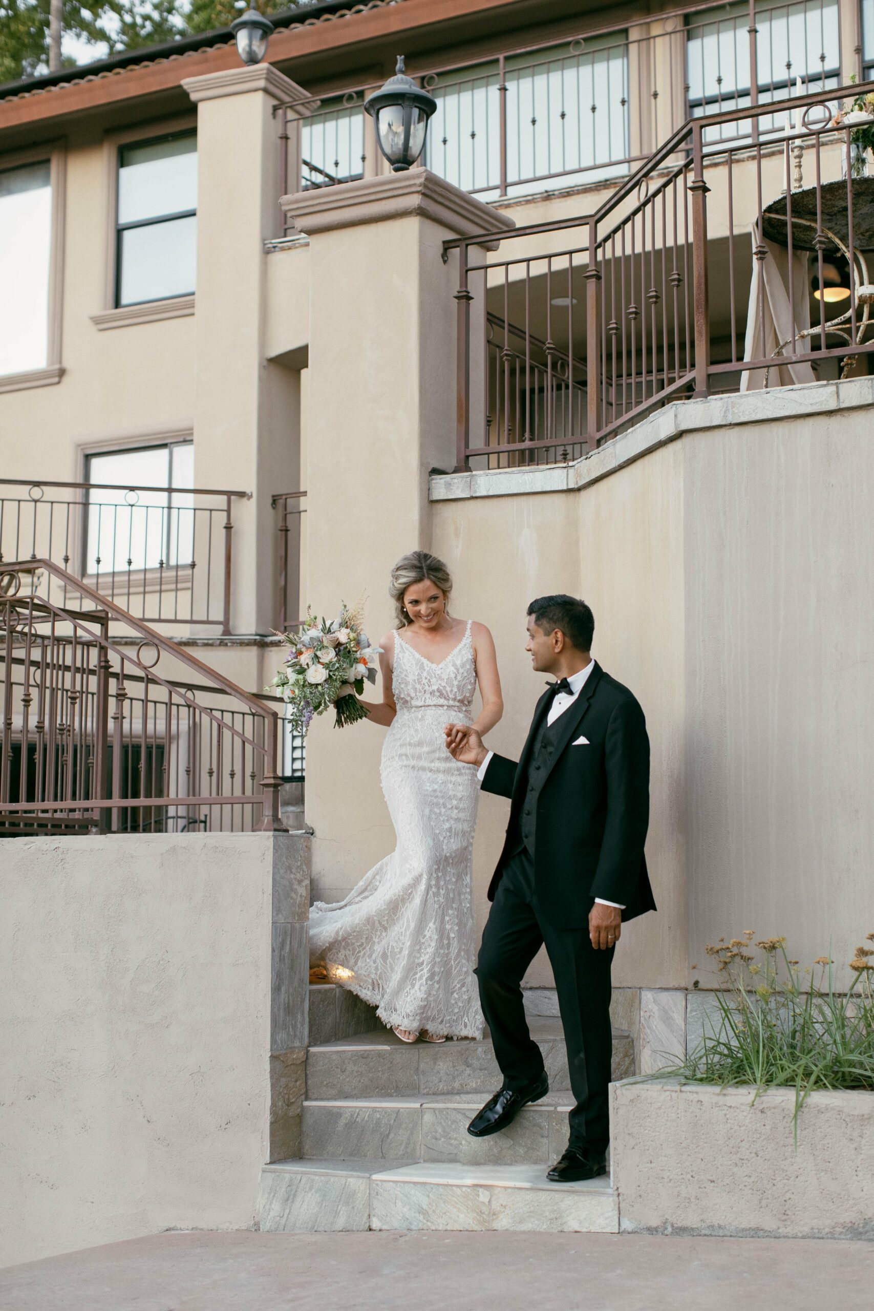 A person in a formal white dress descends stairs, holding a bouquet and being assisted by another person in a black suit. They are outside a multi-story building with a railing.